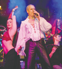 Middle aged white man with balding head wearing a blousy tie-front white shirt with tight shiny dark pants with a woman dancing sideways wearing a red bikini top and low-rider black pants and others dancing behind