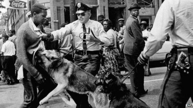 Black and white photo of young black man and white police officer grabbing his shirt in front and a German Shepherd police dog biting at the guy's stomach