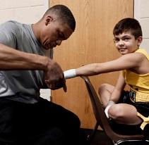 Black man in gray shirt taping up the hand of a young white boy in a yellow tank top