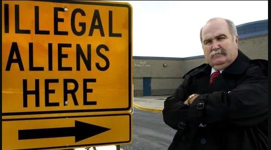 SIgn saying illegal aliens here and a stern bald older man with arms crossed and 