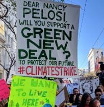 Outdoor rally with signs, one says We Want to Live Here, one says Dear Nancy Pelosi will you support a Green New Deal