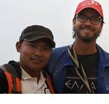 Young looking boy of color with a baseball cap next to a white man with beard and mustache and glasses with a baseball cap on smiling