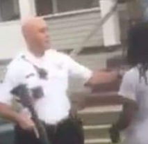 Blurry photo of a white bald policeman in a white shirt and holding a rifle pushing a black man with long braids