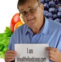 Older white man with glasses in front of fruits and vegetables holding a sign that reads I am healthyfoods.com