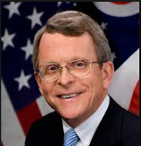 Older white man with combover and wire rimmed glasses smiling in a suit in front of a U.S. flag