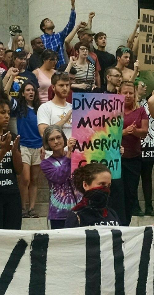 Person holding sign in crowd of people with fists in air, reading Diversity Makes America Great against tie-dyed background
