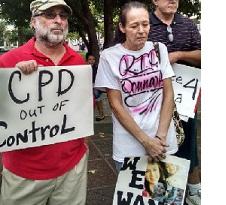 Man with gray beard and sunglasses wearing a baseball cap holding a sign that says CPD out of control standing next to an older woman with gray hair pulled back in a ponytail looking down at the ground, very sad, wearing a white T-shirt with words "RIP Donna" on it and holding a sign with pictures of a young woman on it