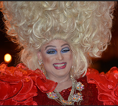Man dressed a woman with huge blonde hair in curls going up in the air lots of blue eye shadow, red lips and a red outfit
