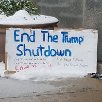 Sign sitting outside in the snow against a large planter says End the Trump Shutdown