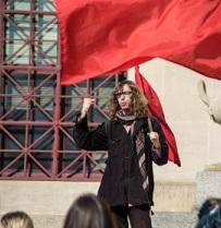 Young white man with long brown curly hair and glasses holding a very large red flag standing in front of a crowd raising his fist in the air