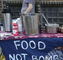 Outside table with pots and pans on it and tablecloth with sign Food not Bombs