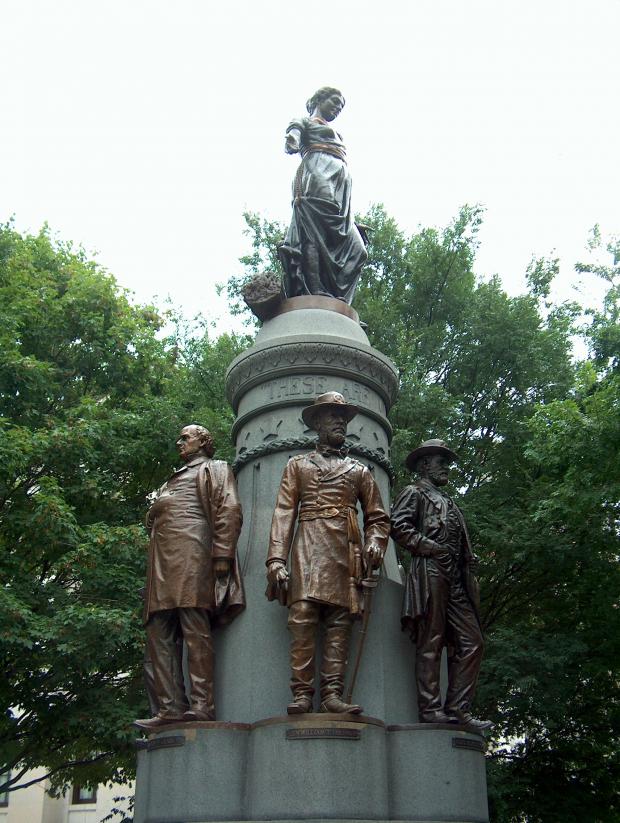 Gray pointy statue with Union Soldier figures standing with their backs around it against trees in the background