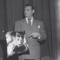 Black and white photo of white man in a suit pointing to a picture of himself in his hand