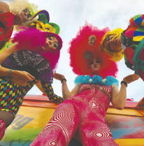 Camera angle looking from ground up at four decked out women with bright orange, pink and rainbow colored fluffy feathery bedazzled outfits
