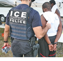 Large white man in uniform that says ICE Police on the back with his back to the camera holding the arm of a dark skinned man in a white t-shirt and shorts with his back to the camera and his hands behind him in handcuffs