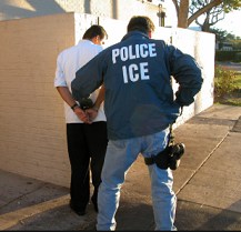 Back of man in blue jacket with white letters reading ICE putting another man with his back to us in handcuffs