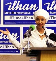 Young woman of color wearing a white hat and suit and pearl necklace standing in front of mics at a podium and signs with her name Ilhan State Representative in the background