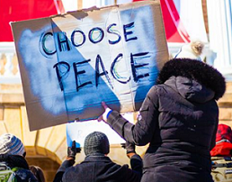 Woman with her back to the camera wearing a large winter coat with furry hood holding a big sign that reads CHOOSE PEACE, she is with a group of people at a rally