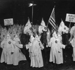 People in white robes with pointy top hats in a circle outside at night with a flag