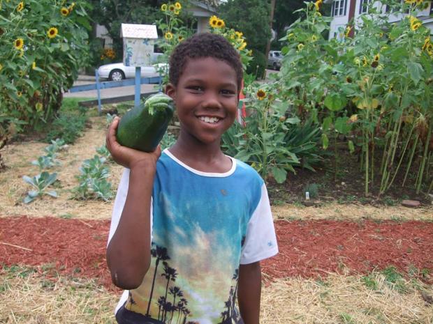 Young black boy smiling and holding a very large zucchini on his shoulder, standing in a garden
