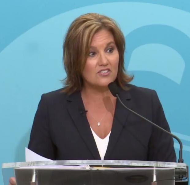 White woman in a black suit with white shirt underneath and necklace with shoulder length brown hair standing at a mic with a blue background, her mouth in a form like she's talking