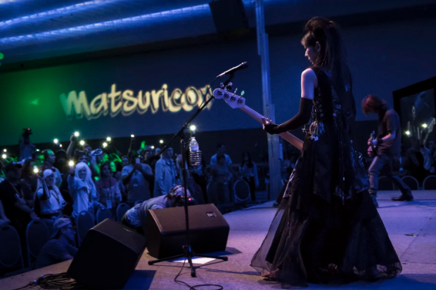 White woman in a Renaissance looking gown playing guitar on stage to a crowd standing under the sign Matsuricon