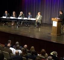 Panel of people on a stage with someone at the podium and an audience