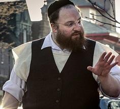 Large white man with jewish hat and wire rimmed glasses, a longish brown beard wearing a white collared button down shirt and black button vest his hand gesturing as he talks to someone to his right