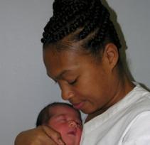 Black woman with hair up in braids side view smiling with eyes looking down as she holds a newborn baby