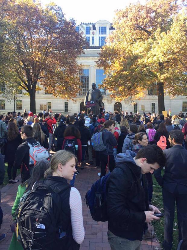 A crowd of people, mostly college students protesting around a statue with two trees on either side and a big building in the background