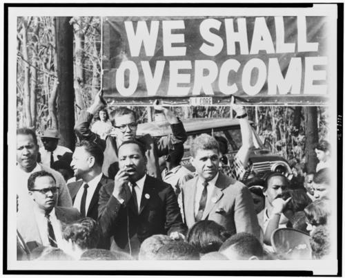 Black and white photo of Martin Luther King Jr. and others marching under sign that says We Shall Overcome