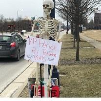 Skeleton leaning against a pole outside holding an anti-Pence sign