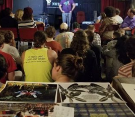 Lots of people with their backs to the camera sitting and watching someone standing at the mic, with art on a table in the foreground