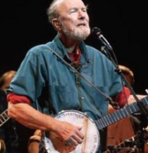 Older gray haired man with beard playing a banjo and singing into a mic