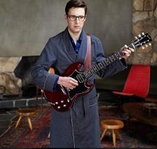 Young geeky guy with big black rimmed glasses wearing a bathrobe holding an electric guitar in a room with two wooden stools