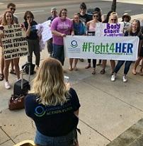 A back of a woman in the foreground speaking outside to a group of women facing her holding signs saying #Fight4Her
