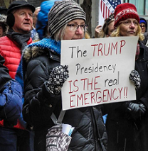 Woman in cold weather gear holding a sign at a protest outside that reads The Trump Presidency is the REAL emergency