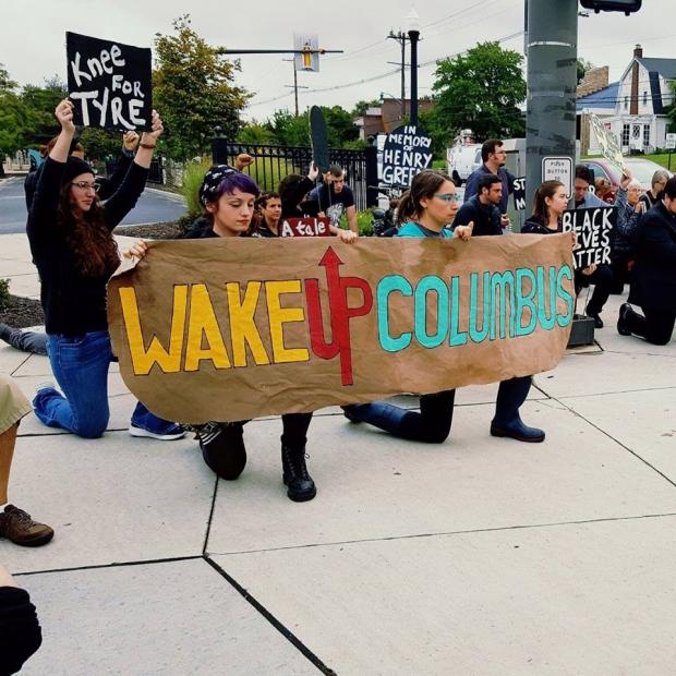 Young white people holding a sign saying Wake Up Columbus kneeling on ground and as part of a rally with lots of people behind them one with a sign saying Knee for Tyre