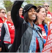 Young black woman with chin length black hair and a cap with her arm in the air and a smile carrying a red sign with white letters about the school system, surrounded by other women and men outside like they are marching