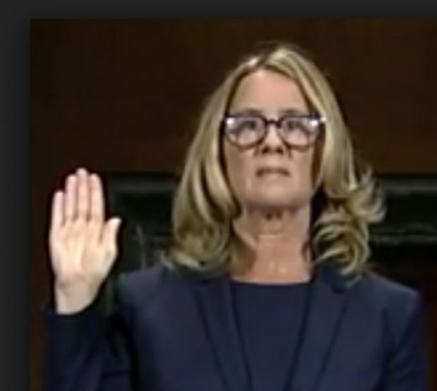 Blonde woman with glasses holding her hand up as if swearing to tell the truth