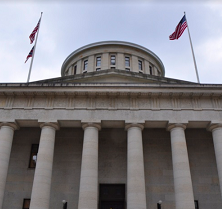 Big white government building with columns and a round top and  flags flying on each side