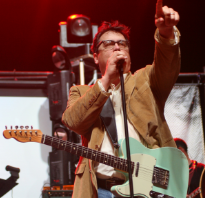 White brwon-haired man with glasses singing at a mic with a guitar around his neck and he's pointing up in the air
