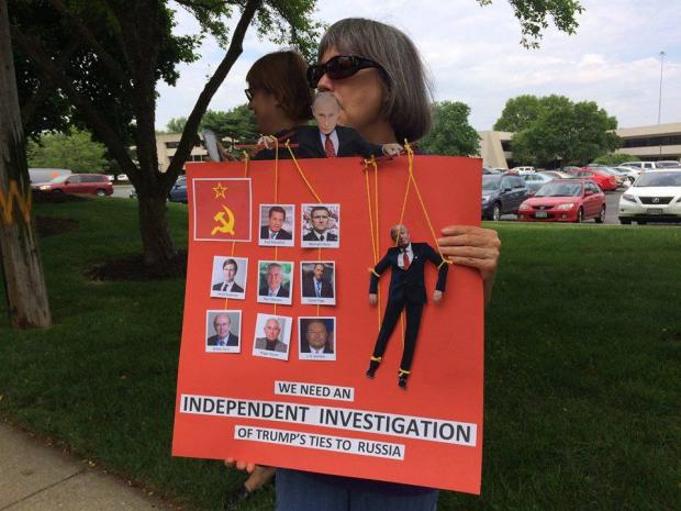 Woman standing outside with sunglasses on holding a huge red sign in front of her asking for an independent investigation of Trump's ties to Russia