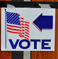 A white sign on a wood wall with a flag and an arrow pointing left and the word VOTE
