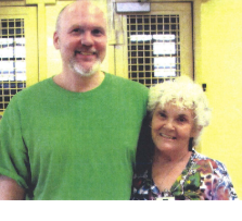 Tall bald white man with a mustache and beard and green T-shirt standing smiling with his arm around a gray haired smiling lady in a flowered dress