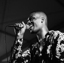 Black and white photo of bald black man smiling and singing into a mic with a flowered shirt on