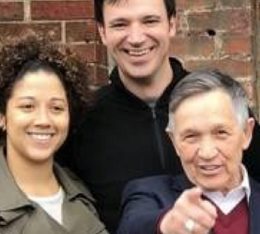 Young black woman smiling on the left, tall white man smiling in the middle and older white man pointing at the camera standing in front of a brick wall