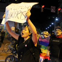 Young woman holding a protest sign above her head shouting and a person behind her in a rainbow flag and a guy next to her in a hat and glasses