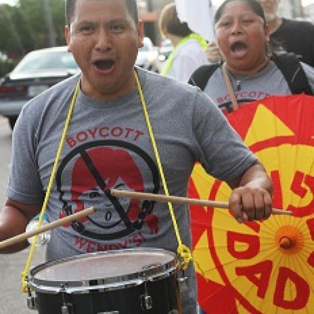 Latino man in foreground with drum around his neck and hitting it with drumsticks, his mouth open chanting, wearing a shirt that says Boycott Wendys with a picture of a girl's head with red pigtails and a No sign around it (circle with line through it) and a Latino woman behind him holding a bright orange and yellow sign also with her mouth open chanting