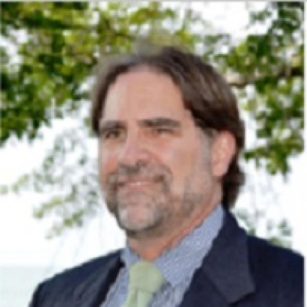 Middle aged white man with short brown hair, a grayish beard and mustache wearing a blue suit standing in front of a tree